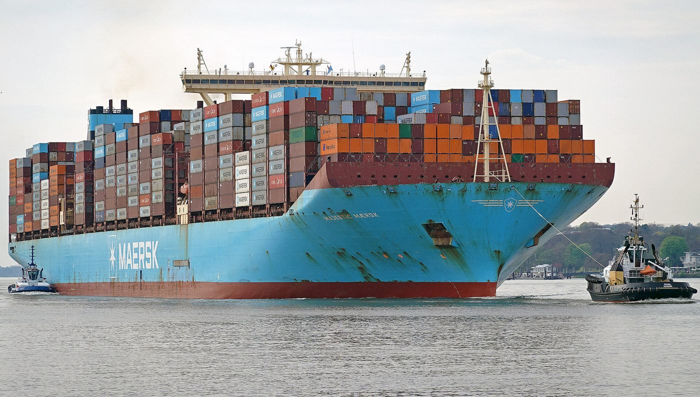 Das blaue Containerschiff Majestic Maersk, voll beladen mit bunten Containern, wird von einem Schlepper begleitet